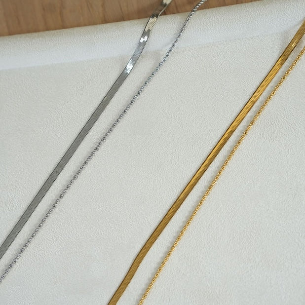 Two gold and silver chains on a white cushion with a wooden surface in the background.
